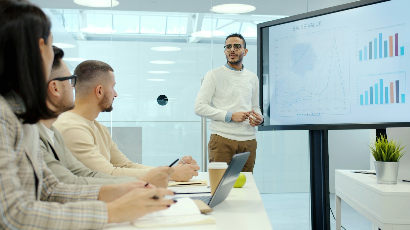 Man presents sales charts to colleagues in a modern office.